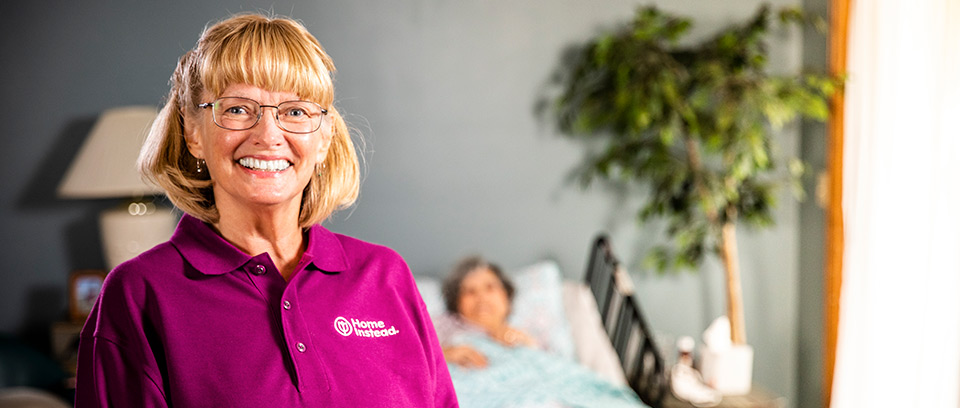 Home Instead Caregiver stands smiling in front of senior woman in bed